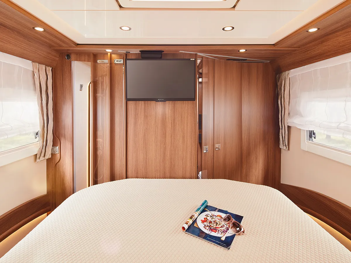 Ein gemütliches Carthago Chic E Line Wohnmobil-Schlafzimmer mit einem ordentlich gemachten Bett, holzgetäfelten Wänden, einem Flachbildfernseher über dem Bett, zwei Fenstern mit durchsichtigen Vorhängen und einer Zeitschrift mit Brille auf dem Bett.