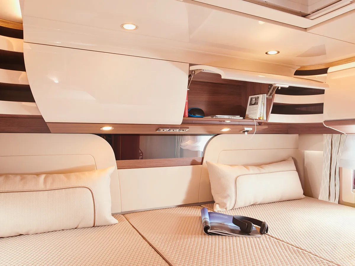 Innenraum eines modernen Carthago Chic E Line Wohnmobil-Schlafzimmers mit beiger Bettwäsche, zwei Kissen, Hängeschränken und einer Zeitschrift mit Sonnenbrille auf dem Bett. Warme Beleuchtung und schlichte Holzakzente schaffen eine gemütliche Atmosphäre.