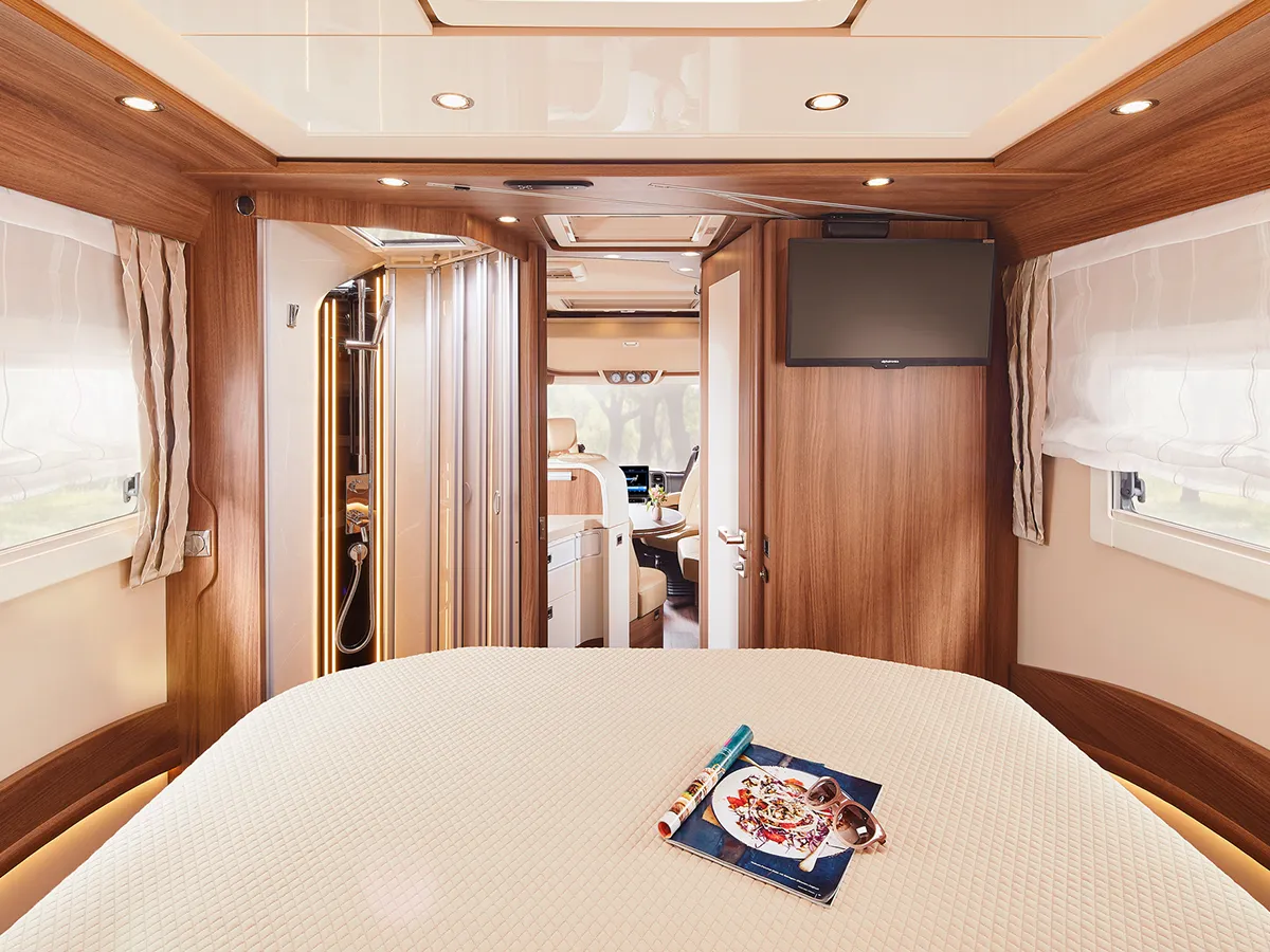 Innenraum eines modernen Carthago Chic E Line Wohnmobil-Schlafzimmers mit Holzvertäfelung, einem großen Bett, Zeitschriften auf der Decke, einem an der Wand montierten Fernseher, Fenstern mit Vorhängen und einem Blick durch offene Türen in die Küche und den Fahrerbereich.