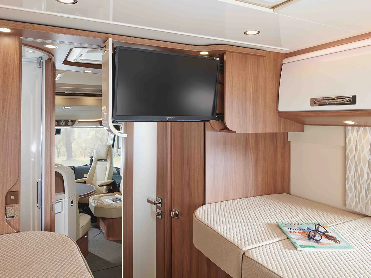 Innenraum eines modernen Carthago Chic E Line Wohnmobils mit holzvertäfelten Wänden, einem montierten Flachbildfernseher, gepolsterten Betten und einer Zeitschrift auf dem Tisch. Sitze und Fenster im vorderen Bereich schaffen einen gemütlichen, funktionalen Wohnraum.