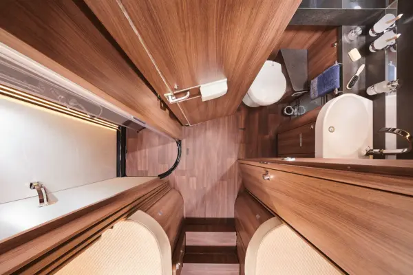 Draufsicht auf ein modernes Carthago Chic S Plus Bad mit Holzverkleidung, Toilette, Waschbecken, Handtuch, Glas, Seifenspender und Spiegelflächen, die einen kompakten und stilvollen Raum schaffen.