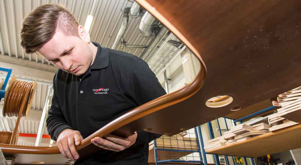 Une personne vêtue d’une chemise noire examine une plaque de bois courbée dans un atelier rempli de pièces en bois et éclairé par des lampes industrielles.