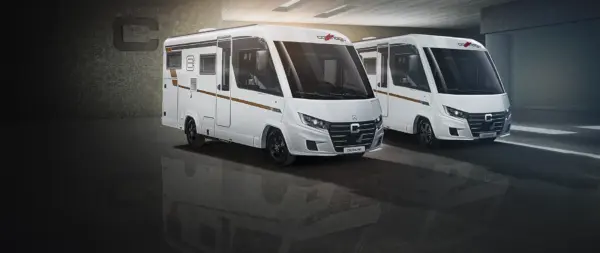 Two modern white motorhomes, including a Carthago C2 Tourer with a sleek design, are parked side by side in an interior setting and are brightly lit to showcase their exterior features and branding.