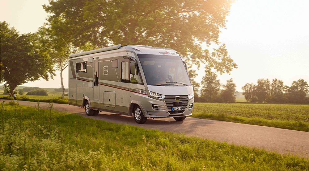 An Iveco Daily motorhome travelling down a narrow country road, surrounded by green fields and trees. The sunlight filtering through the sky creates a warm, serene atmosphere.