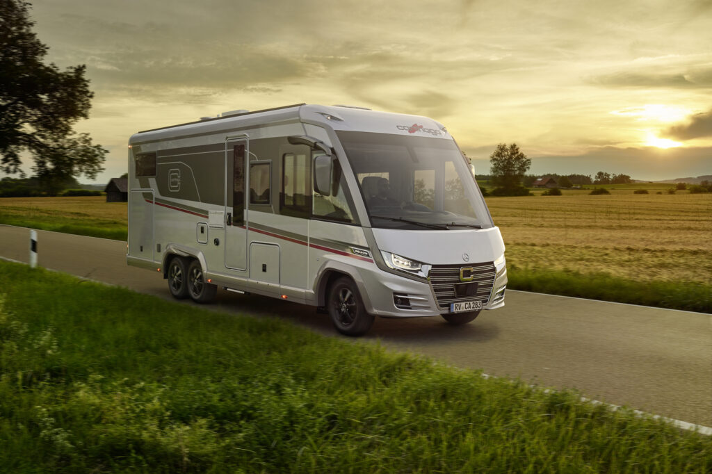 Ein modernes weiß-graues Wohnmobil fährt bei Sonnenuntergang eine Landstraße entlang, umgeben von grünem Gras und Feldern, mit Bäumen und fernen Häusern im Hintergrund.