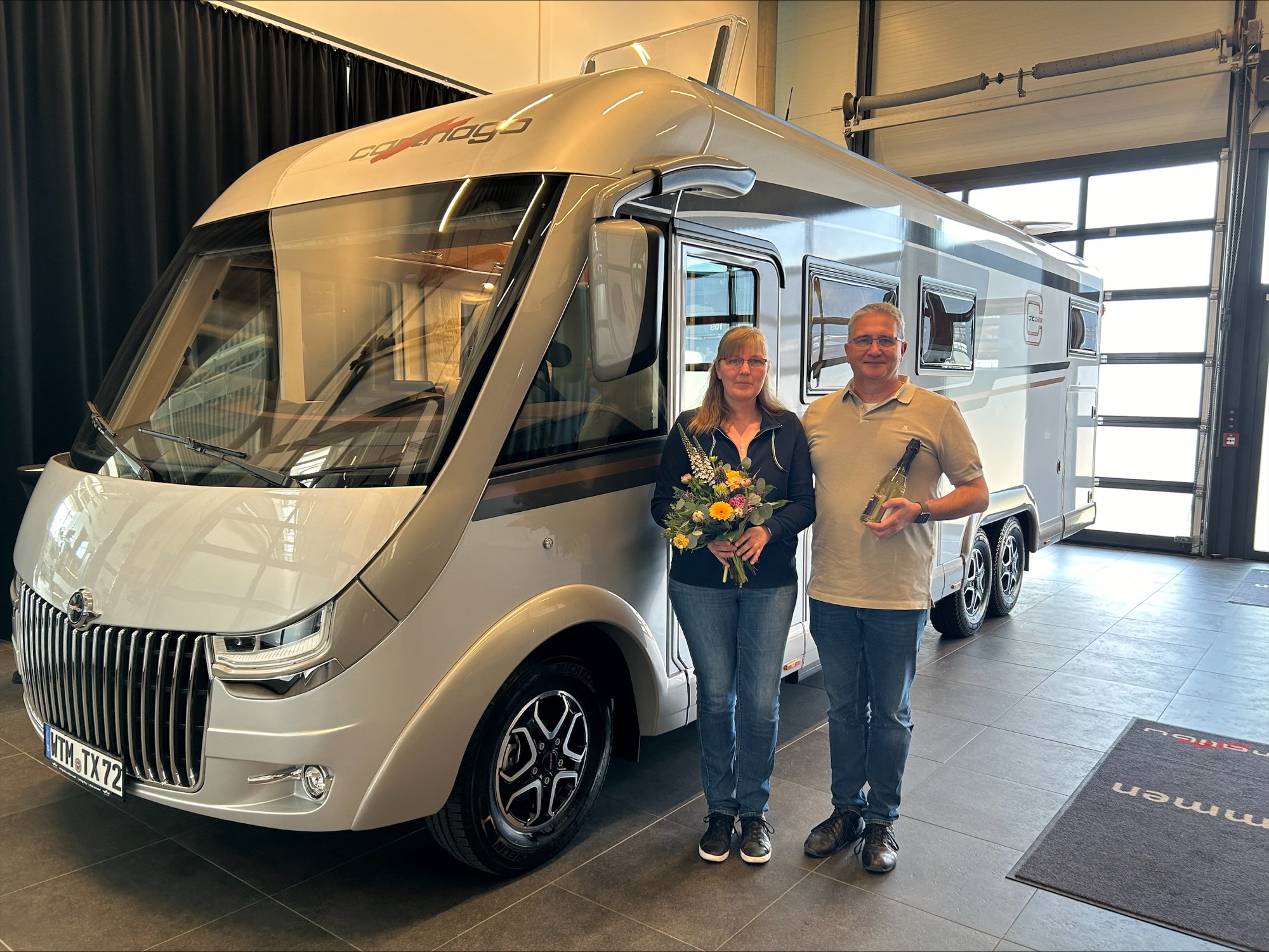 Ein lächelnder Mann und eine lächelnde Frau stehen drinnen vor einem großen weißen Wohnmobil beim Carthago Werksverkauf; die Frau hält einen Blumenstrauß in der Hand und der Mann zeigt stolz eine kleine Trophäe.