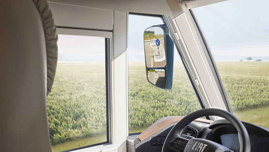 Uitzicht vanuit het interieur van een Fiat-voertuig met stuurwiel, zijraam en zijspiegel, waarin een verkeersbord wordt weerspiegeld. Buiten zijn een groen grasveld en een open hemel te zien.