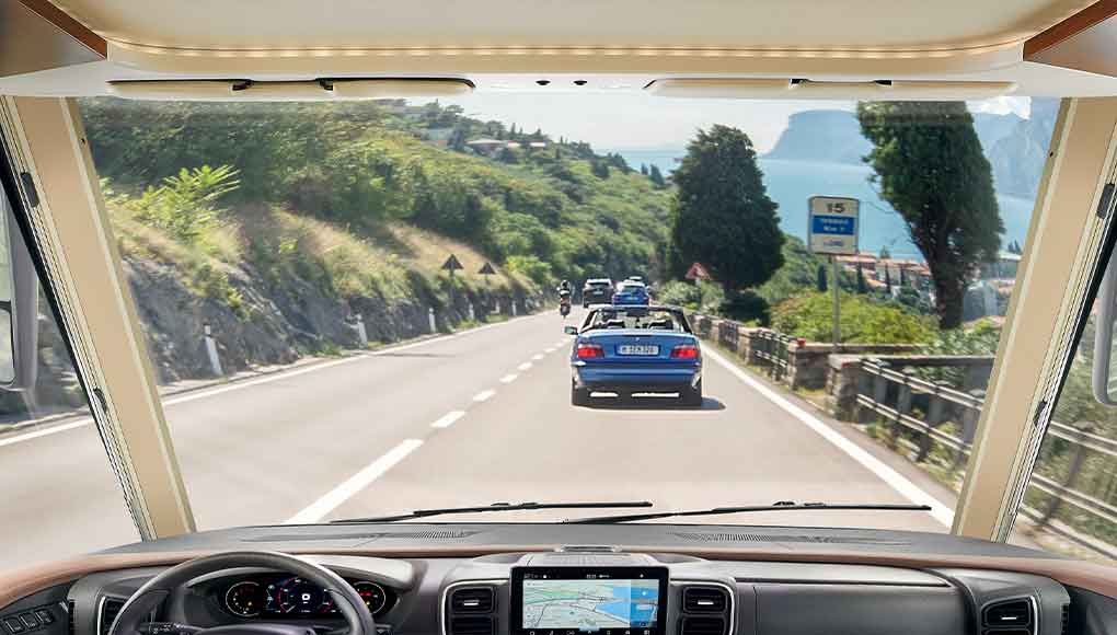 Uitzicht vanuit het interieur van een voertuig door de voorruit op een schilderachtige, bochtige weg, omzoomd door bomen en vangrails, waarop meerdere auto’s rijden en in de verte een meer te zien is.