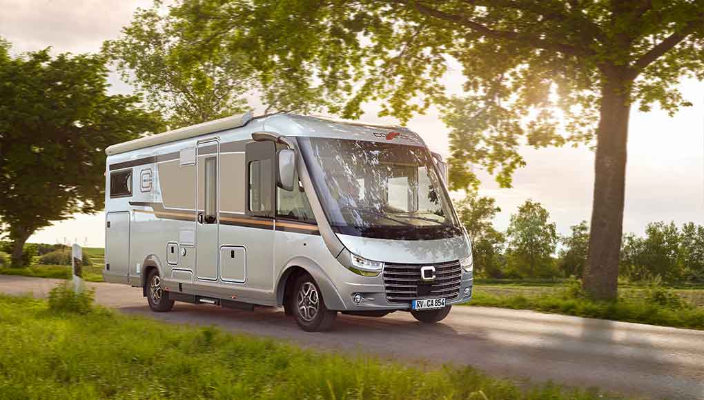 Een zilverkleurige camper rijdt over een landweg die wordt omzoomd door groen gras en bomen, terwijl het zonlicht door de takken schijnt.
