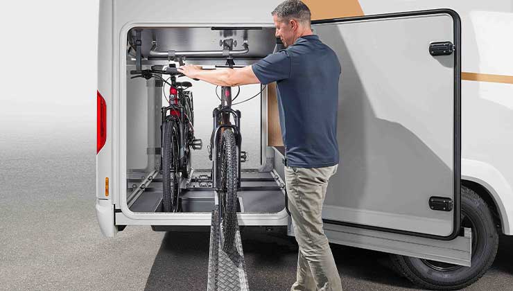 A man is loading two bikes into the rear storage space in a motorhome, using the built-in sliding bike rack for easy access and secure transport.