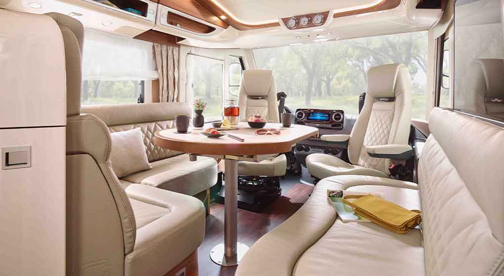 L’intérieur d’un camping-car moderne, avec des sièges en cuir couleur crème, une table ronde en bois sur laquelle sont disposées des boissons et des collations, de grandes baies laissant entrer la lumière du jour et offrant une vue sur la végétation luxuriante à l’extérieur.
