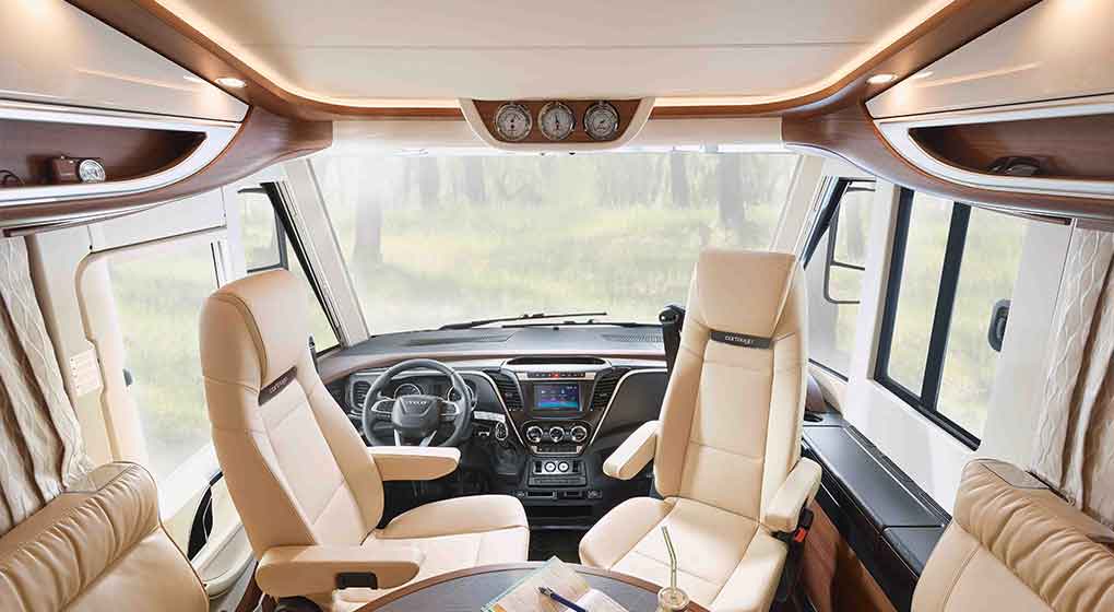 L’intérieur d’un camping-car moderne, avec deux fauteuils en cuir marron clair, une petite table ronde et un grand pare-brise donnant sur un paysage extérieur flou parsemé de plantes vertes. Le tableau de bord est équipé de commandes numériques et rehaussé de touches de bois.