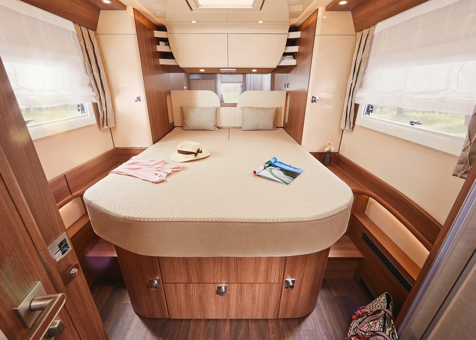 Ein gemütliches, helles Schlafzimmer in einem Carthago Chic S Plus Wohnmobil mit einem großen Bett mit Kissen, einem Hut und Büchern darauf, Holzvertäfelung, Fenstern mit Vorhängen auf beiden Seiten und Stauschubladen unter dem Bett.