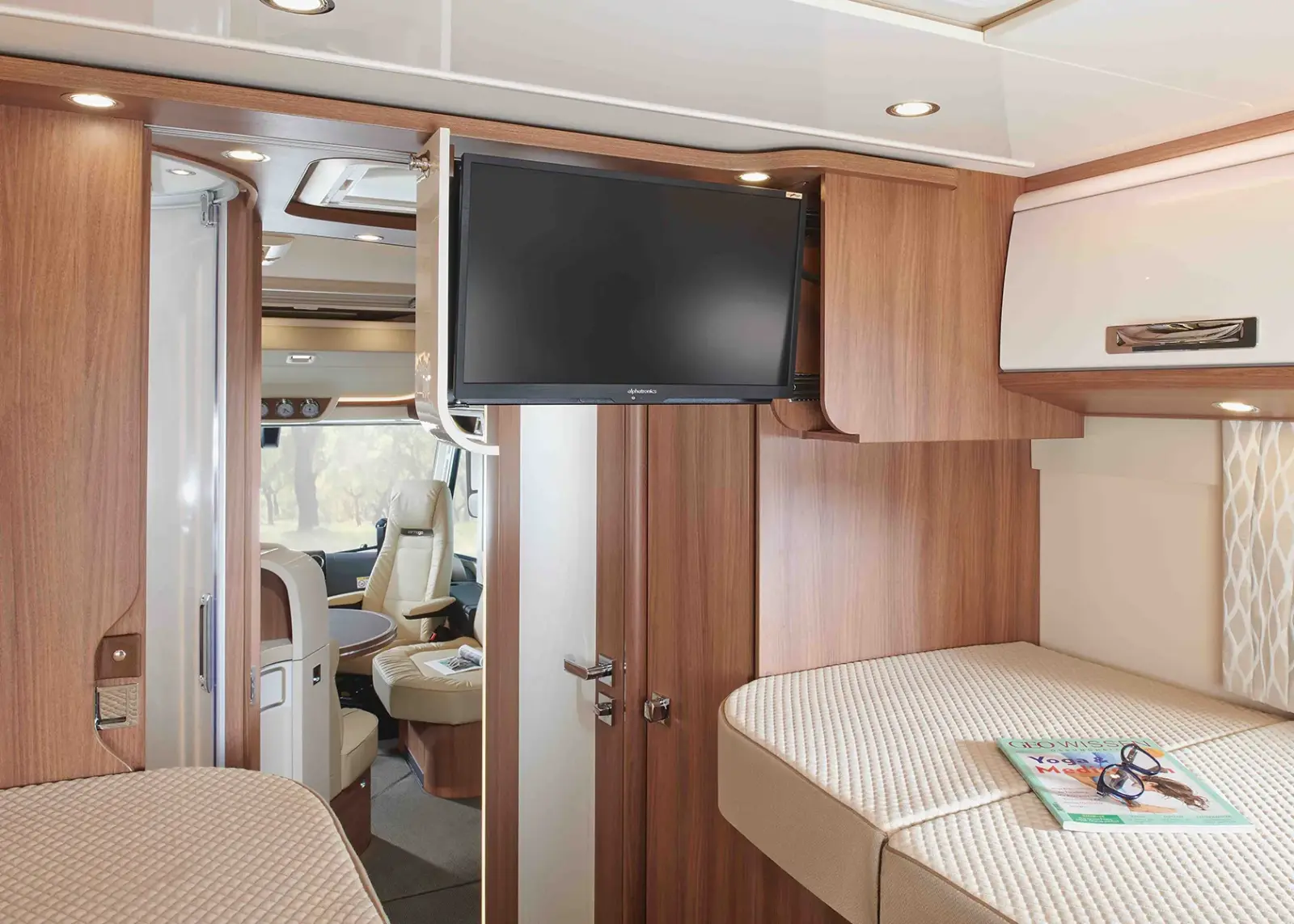 Innenraum eines modernen Carthago Chic S Plus Wohnmobils mit Holzvertäfelung, einem montierten Flachbildfernseher, beigen Sitzen und Betten sowie einer Zeitschrift mit Brille auf einem Bett. Durch die Fenster strömt Licht herein und schafft eine gemütliche Atmosphäre.