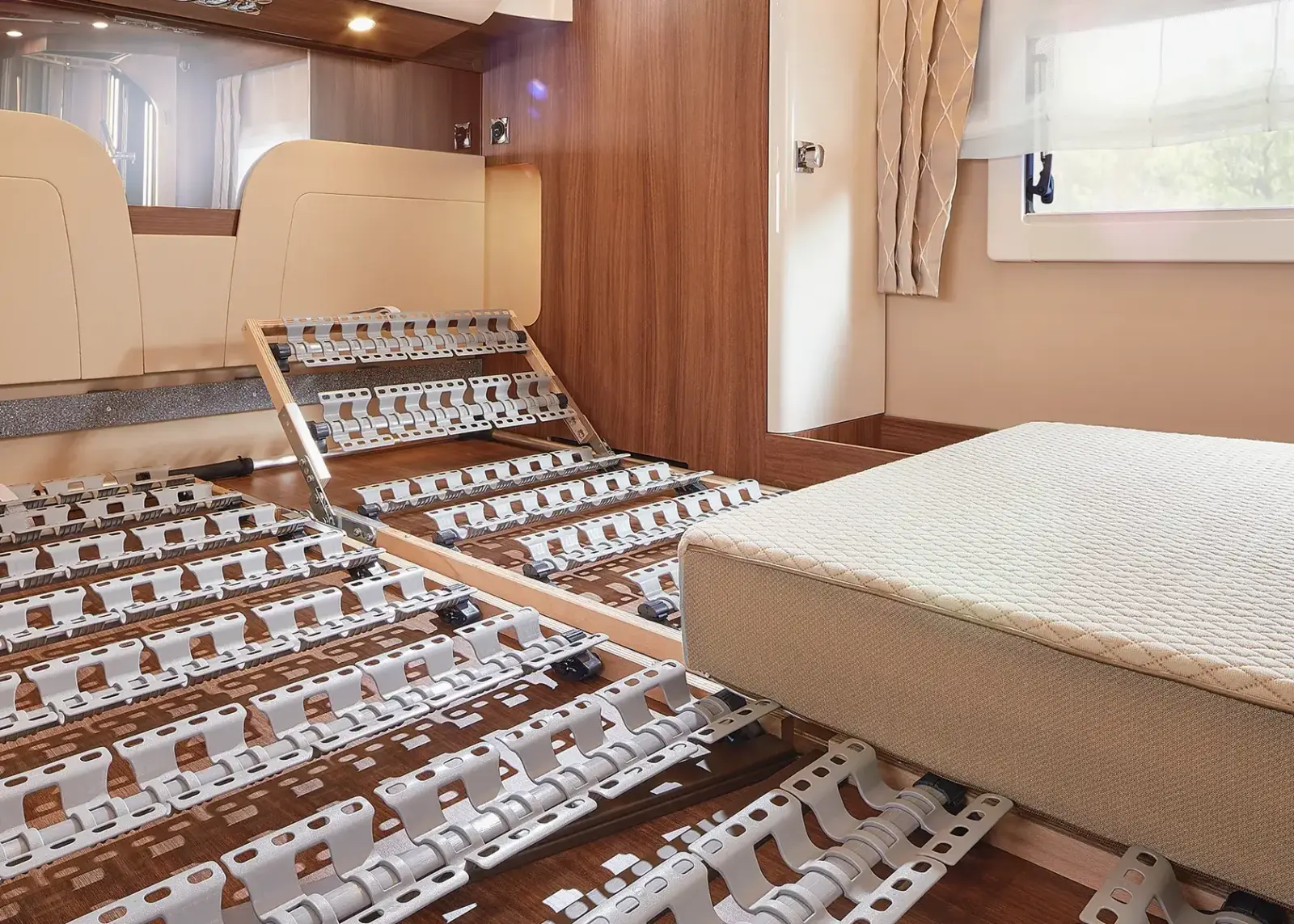 Ein Zimmer mit Holzfußboden und einem ungewöhnlichen Bettgestell aus mehreren verstellbaren Kunststofflatten, von denen ein Teil hochgeklappt ist, was an die innovative Innenausstattung eines Carthago Chic S Plus erinnert, sowie einer Matratze, die in der Nähe eines Fensters mit beigen Vorhängen liegt.