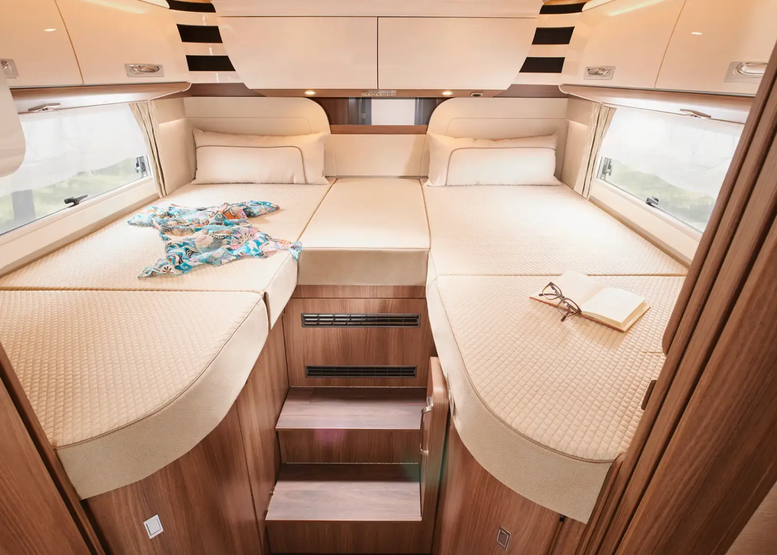 Ein gemütliches Carthago Chic S Plus Wohnmobil-Schlafzimmer mit drei beigen Matratzen, die eine große Schlaffläche bilden, Holzstufen in der Mitte, ein offenes Buch mit Brille und ein gefaltetes buntes Hemd auf dem Bett, umgeben von Holzschränken und hellen Fenstern.