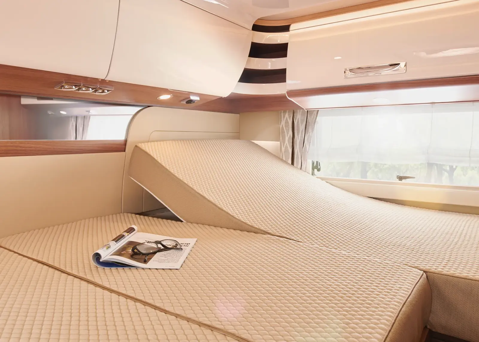 Ein gemütliches Carthago Chic S Plus Wohnmobil-Schlafzimmer mit einem beigen, verstellbaren Bett mit geteilter Matratze. Eine Seite ist erhöht, und auf dem Bett liegt eine Zeitschrift mit Lesebrille. Hängeschränke und ein großes Fenster füllen den Raum mit natürlichem Licht.