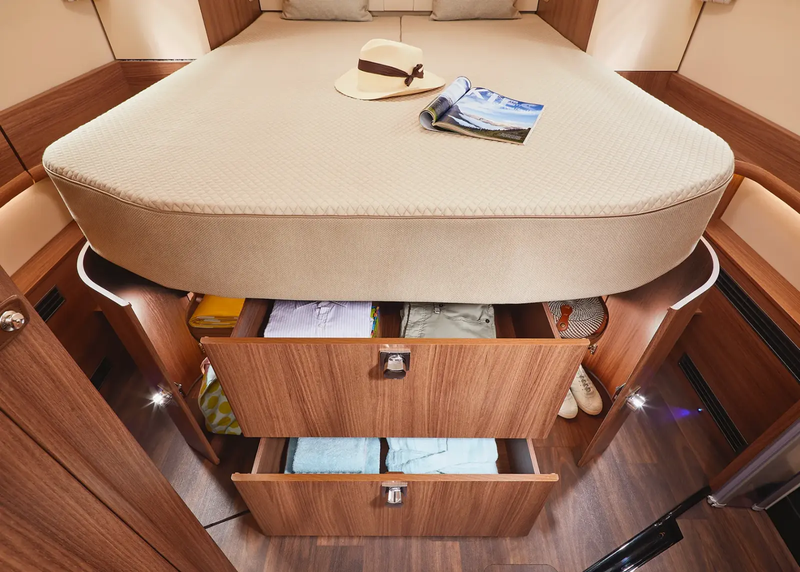 Ein ordentlich gemachtes Bett mit einem Hut und einer Zeitschrift darauf, erhöht über zwei offenen Holzschubladen, die mit Kleidung gefüllt sind, in einem gemütlichen, modernen Carthago Chic S Plus Zimmer mit Holzfußboden und warmer Beleuchtung.