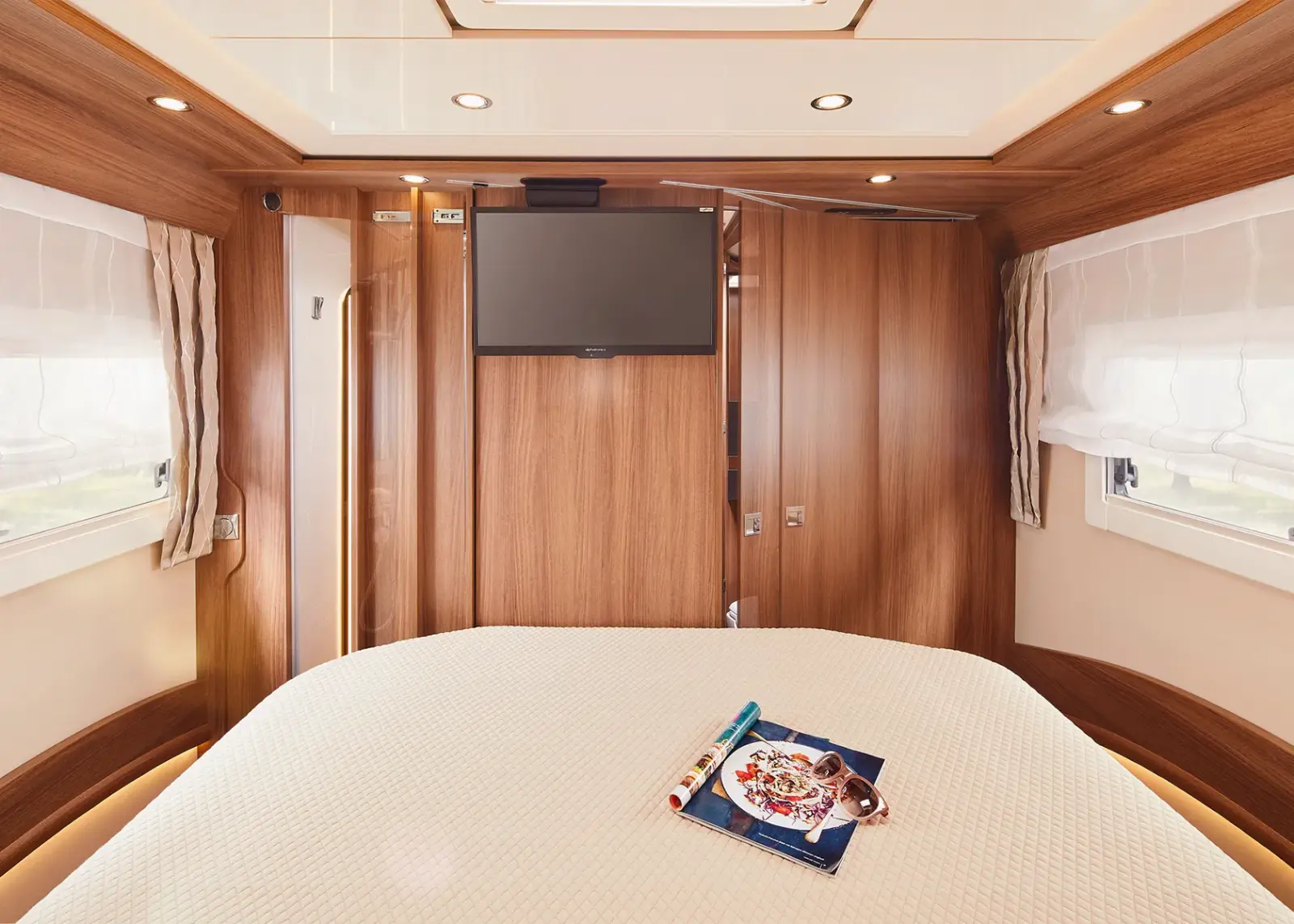 Ein gemütliches Schlafzimmer in einem Carthago Chic S Plus Wohnmobil mit holzgetäfelten Wänden, einem großen Bett mit weißem Strukturbezug, einem an der Wand montierten Fernseher und einer Zeitschrift mit Brille und Stift. Zwei Fenster mit weißen Vorhängen lassen viel Licht herein.