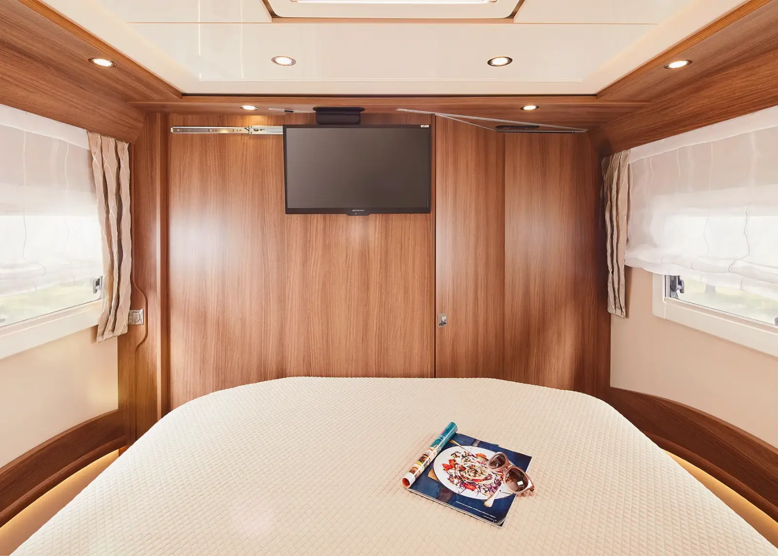 Ein gemütliches Carthago Chic S Plus Wohnmobil-Schlafzimmer mit einem großen Bett, das mit einer weißen, strukturierten Decke bezogen ist, holzgetäfelten Wänden, einem an der Wand montierten Fernseher und zwei Fenstern mit transparenten Vorhängen. Eine Zeitschrift und eine Brille liegen einladend auf dem Bett.