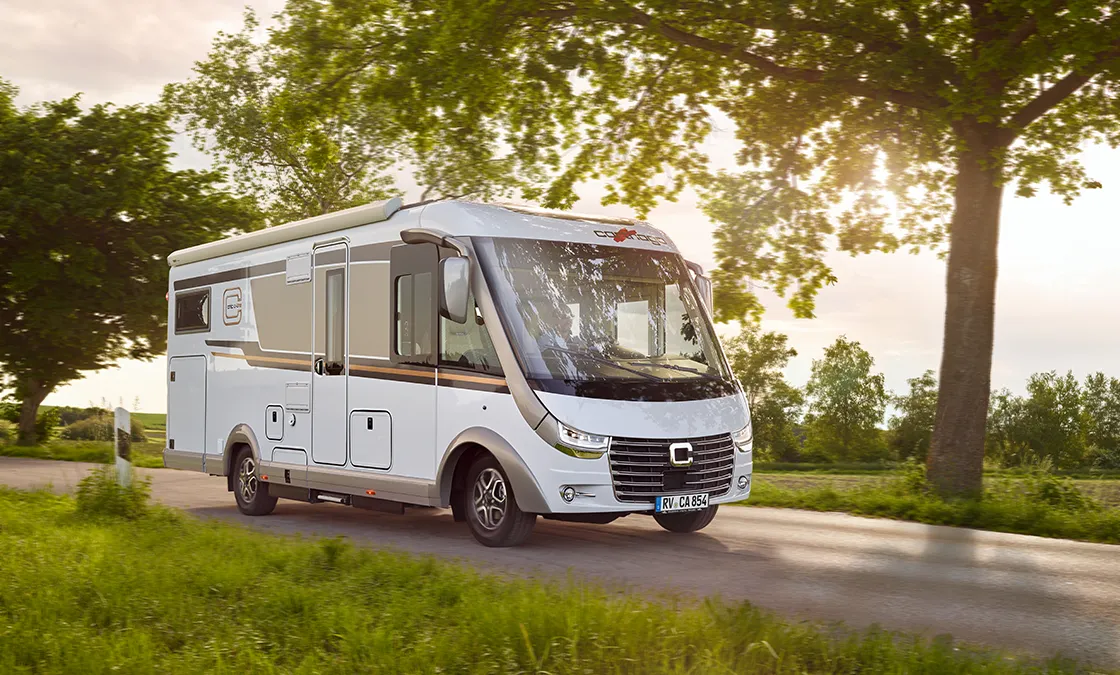 Ein weißes Wohnmobil fährt eine Landstraße entlang, die von grünem Gras und Bäumen umgeben ist, wobei das Sonnenlicht durch die Blätter fällt und der Himmel im Hintergrund klar ist.