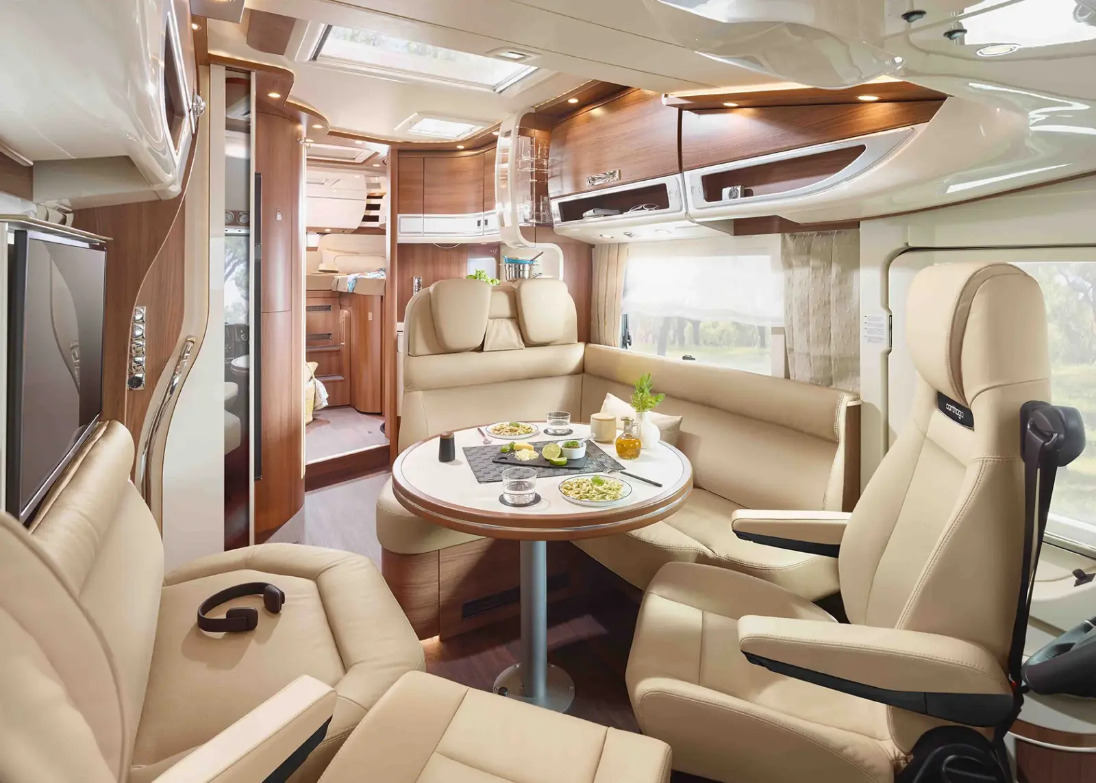 Modernes Wohnmobil-Interieur mit beigen Ledersitzen, einem runden Esstisch mit Speisen, hellen Holzakzenten und viel Tageslicht durch große Fenster. Das Design des Carthago Chic S Plus bietet einen komfortablen und einladenden Raum für entspanntes Reisen.