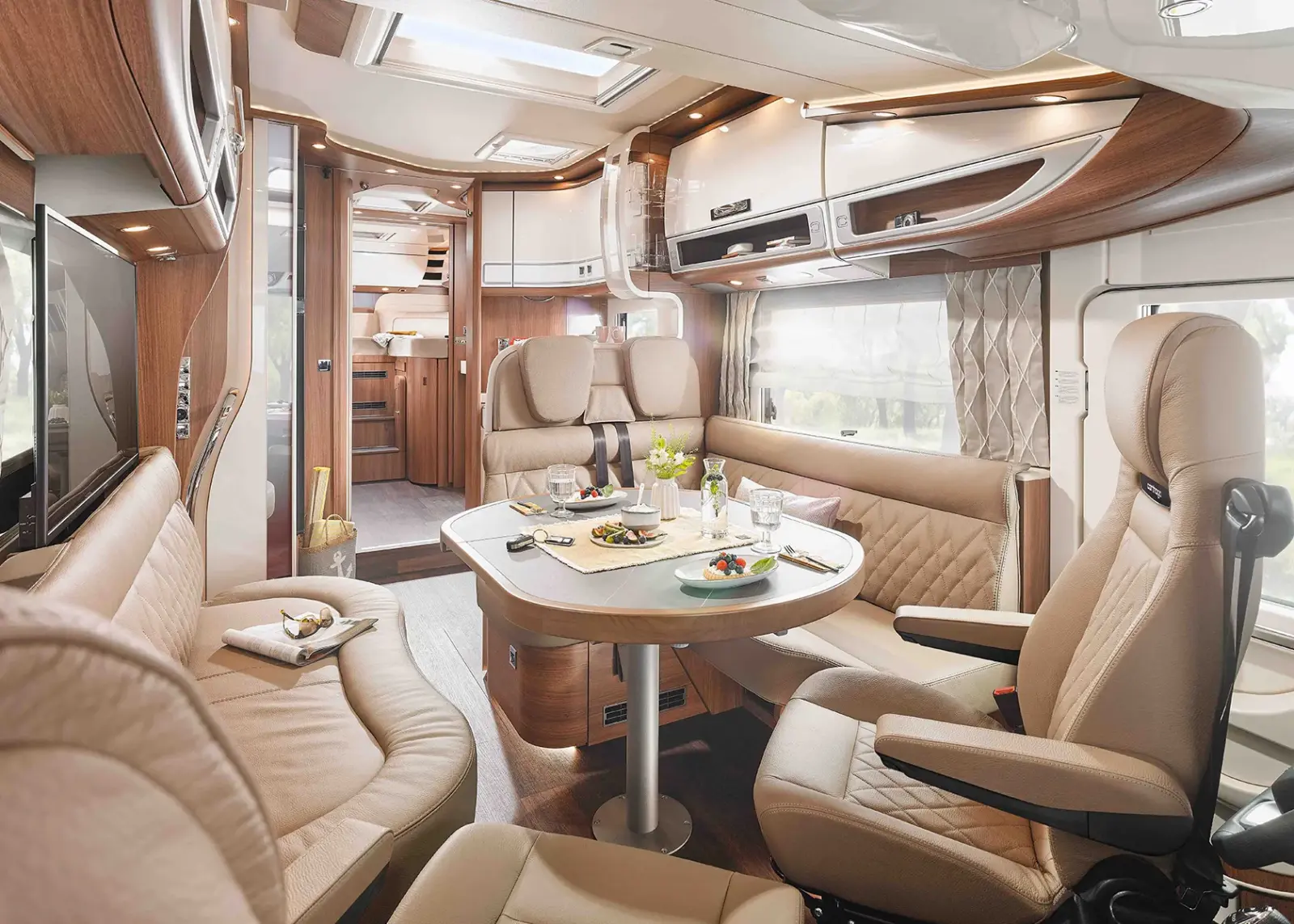 Luxuriöses Carthago Chic S Plus Wohnmobil-Interieur mit beigen Ledersitzen, einem Esstisch mit Tellern und Gläsern, einem geschwungenen Sofa, Holzschränken und großen Fenstern. Der Raum wirkt geräumig und modern.