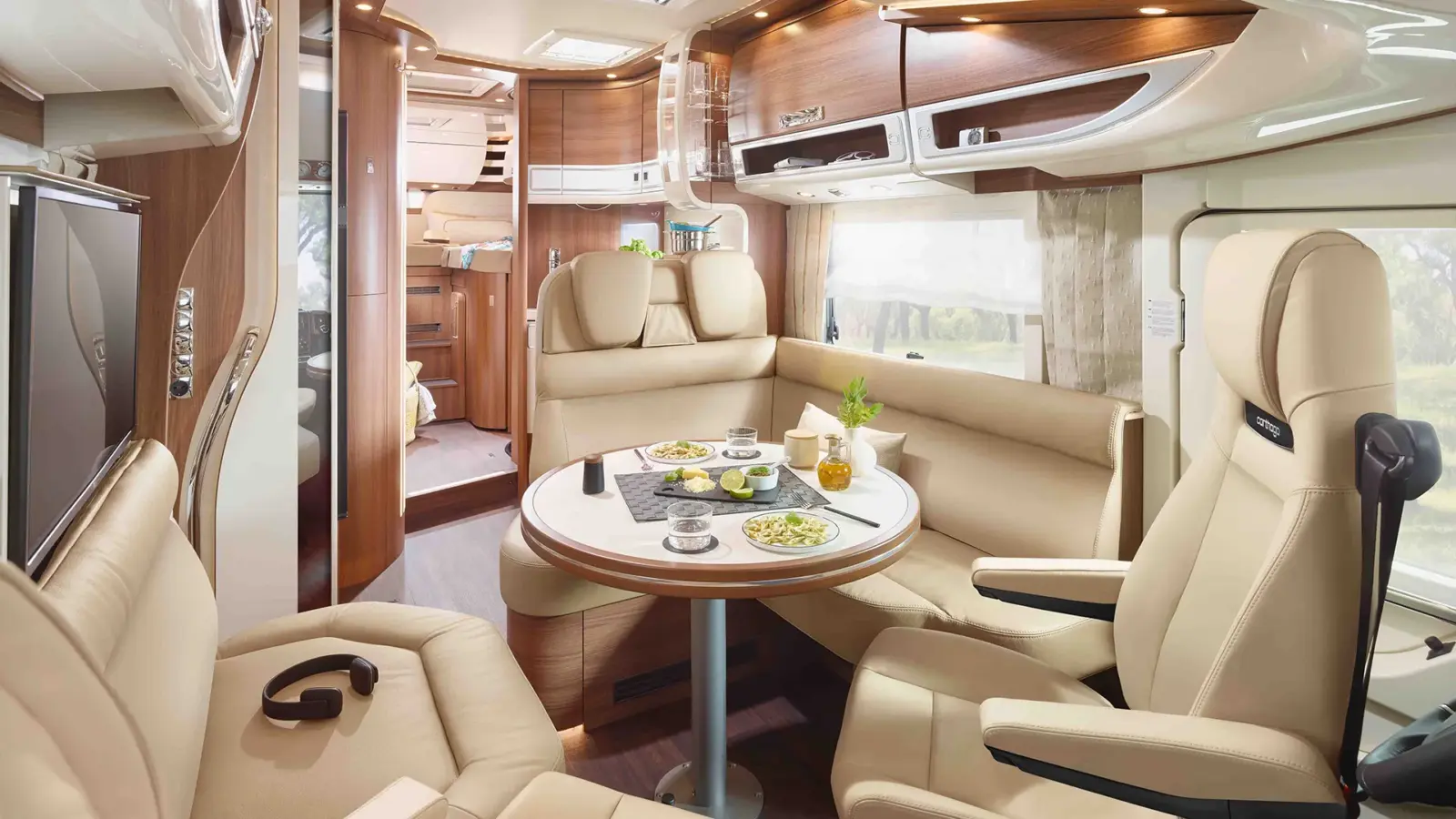Modernes Carthago Chic E Line Wohnmobil-Interieur mit beigen Ledersitzen, einem runden Esstisch mit Tellern, Geschirr und Getränken, Holzschränken und einer gemütlichen, gut beleuchteten Atmosphäre. Im Hintergrund sind ein Bett und ein Küchenbereich zu sehen.