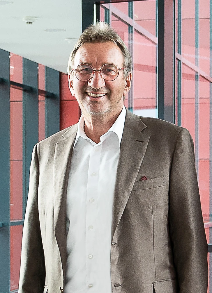 A smiling man with glasses, a beige suit and a white shirt stands in an interior in front of large windows with red and black panelling in the background.
