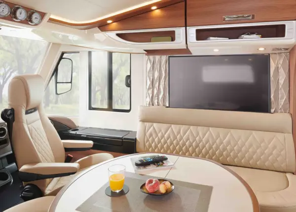 Modernes Wohnmobil-Interieur mit cremefarbenen, gesteppten Sitzen, einem großen Flachbildfernseher und einem runden Tisch für das Frühstück. Große Fenster füllen diesen Carthago Chic E Line Raum mit natürlichem Licht und Blick auf die Bäume draußen.