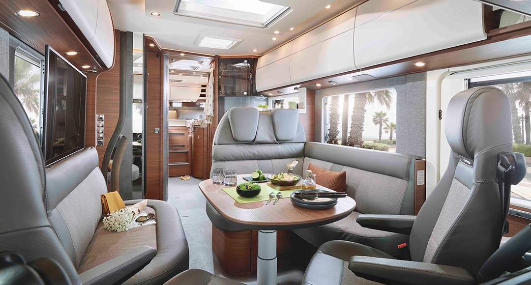 Gli interni di un moderno camper Carthago Chic C Line presentano eleganti superfici in legno, un tavolo da pranzo con piatti e bicchieri, sedute imbottite, una grande finestra con vista sulle palme e tanti vani di stivaggio nella parte alta.