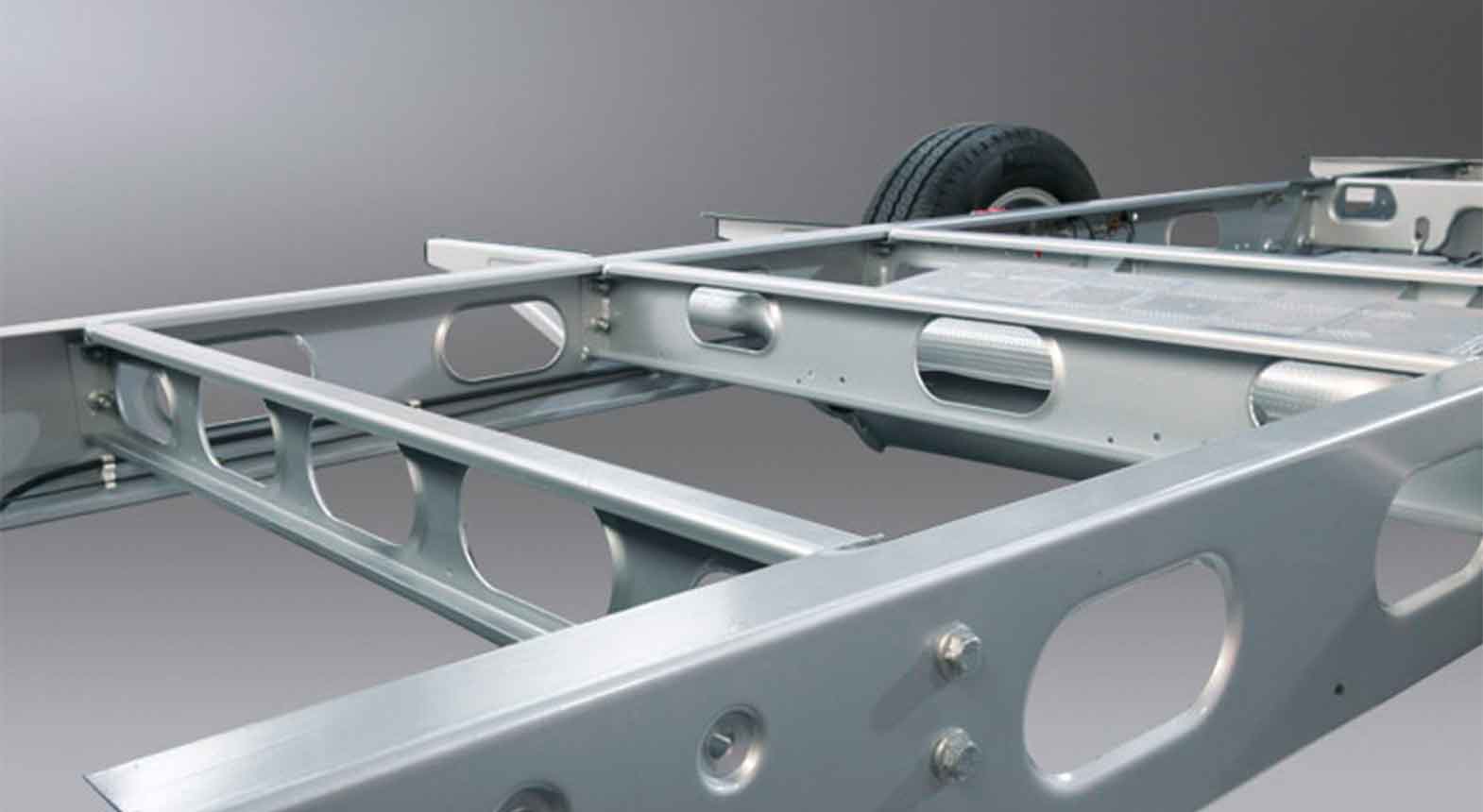 Nahaufnahme eines Fahrgestellrahmens aus Metall, ideal für wohnmobil gewichtskontrolle, mit einem einzelnen Reifen im Hintergrund. Strukturelle Balken und Querträger sind vor einem Hintergrund mit grauem Farbverlauf zu sehen.
