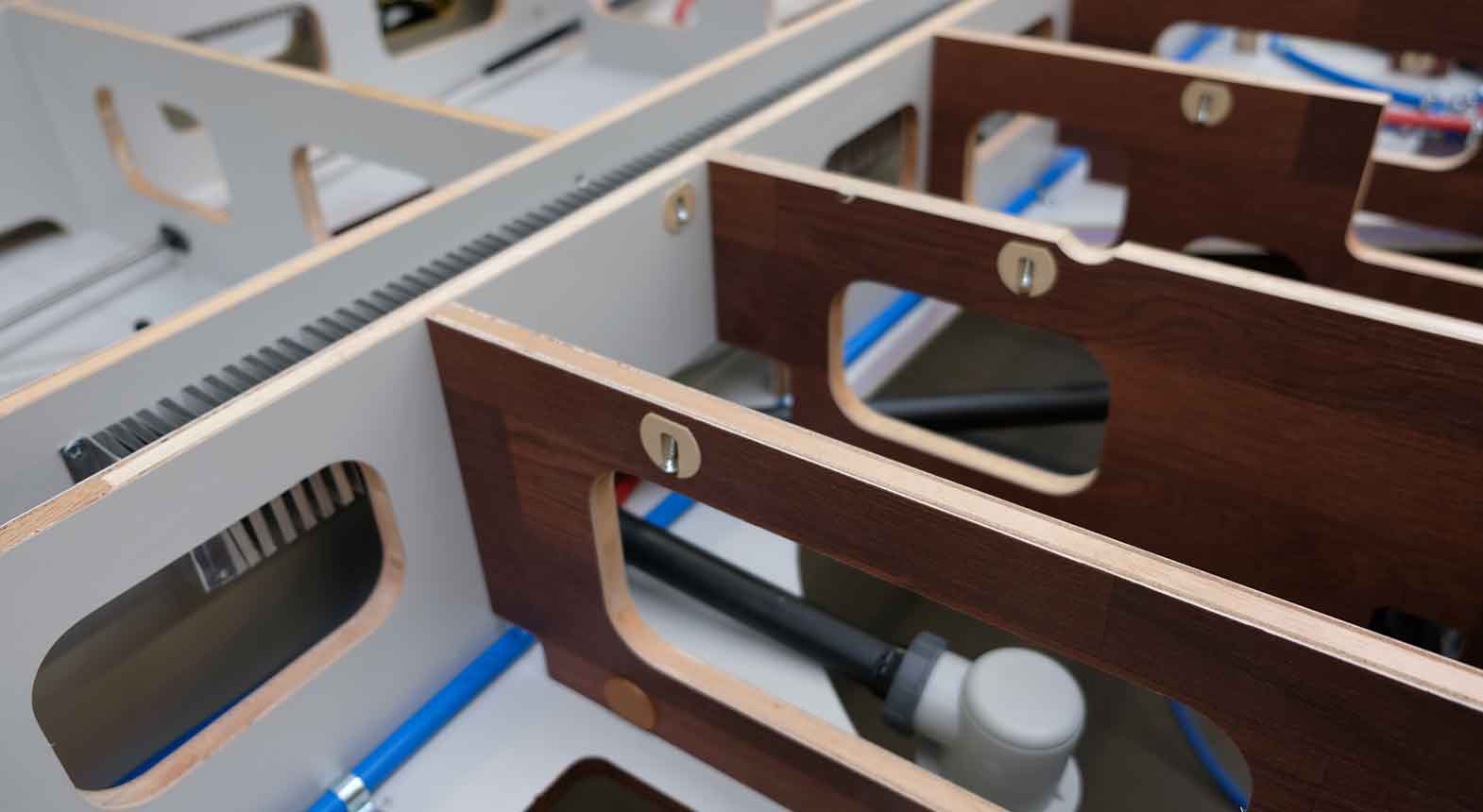 Nahaufnahme einer Anordnung von Holzplatten mit Ausschnitten, die blaue und schwarze Rohre - möglicherweise für ein wohnmobil gewichtskontrollsystem - zeigen, die unter und durch die Struktur verlaufen, Teil des Innenausbaus oder der Möbelmontage.