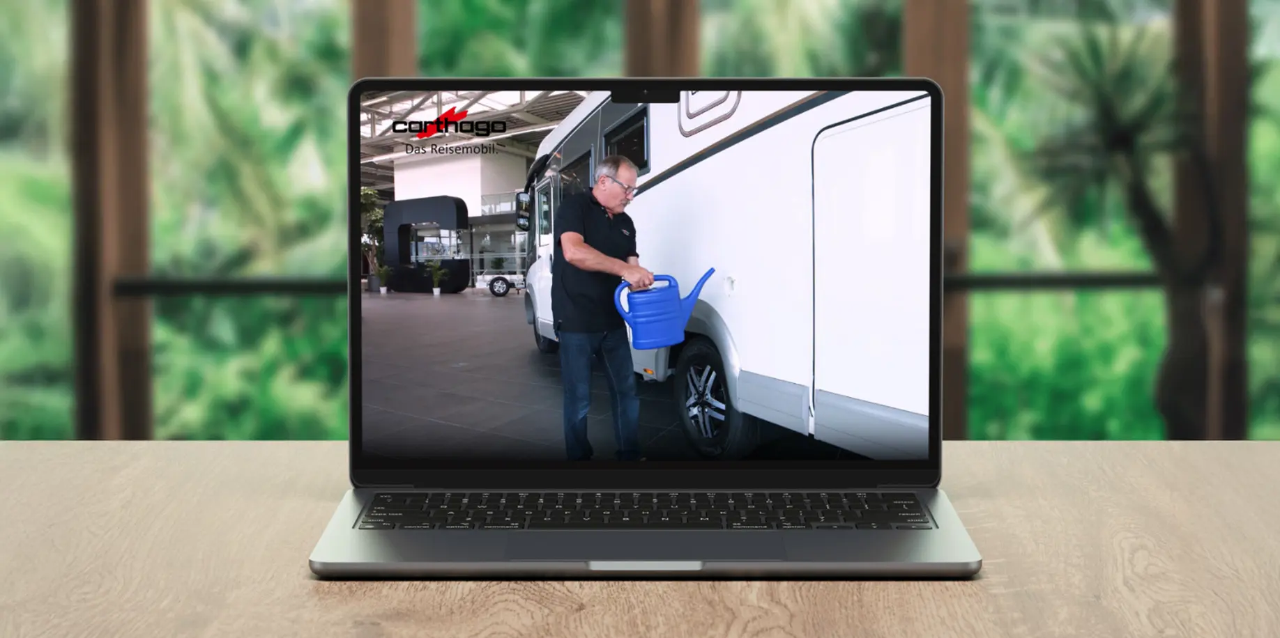 Auf einem Laptop auf einem Holztisch ist ein Video zu sehen, das eine Person zeigt, die neben einem weißen Wohnmobil steht und eine blaue Gießkanne neben dem Lenkrad hält. Im Hintergrund ist unscharfes Grün zu sehen.