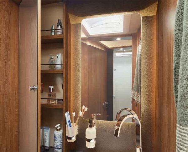 Das kleine Badezimmer im Carthago C Tourer Teilintegriert verfügt über Holzschränke, einen großen Spiegel, offene Regale mit Toilettenartikeln, ein rechts hängendes Handtuch und ein modernes Waschbecken mit Zahnbürstenhaltern auf dem Tresen.