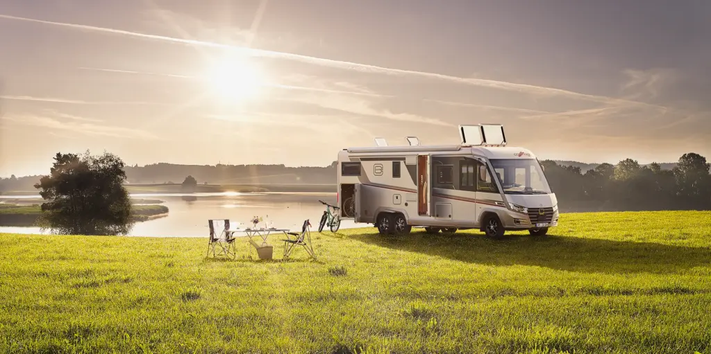 Ein Wohnmobil ist bei Sonnenuntergang auf einer Wiese in der Nähe eines Sees geparkt. Klappstühle, ein Tisch und ein Fahrrad sind draußen aufgestellt, und das Sonnenlicht wirft einen warmen Schein auf die friedliche Landschaft.
