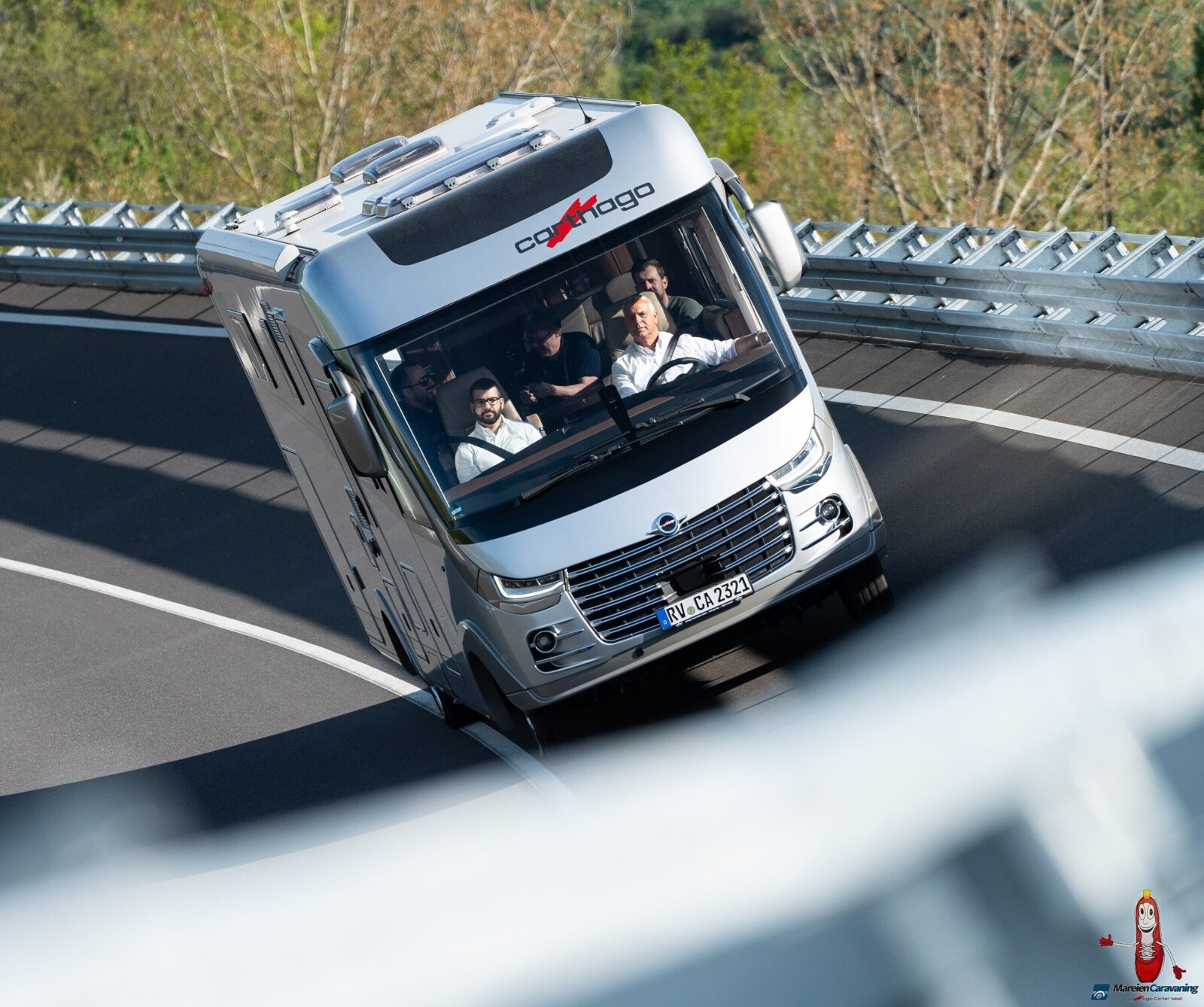 Ein silbernes Wohnmobil mit fünf Personen im Inneren kippt während der Fahrt auf einer kurvigen, geneigten Straße mit Metallgeländern und Bäumen im Hintergrund stark um.