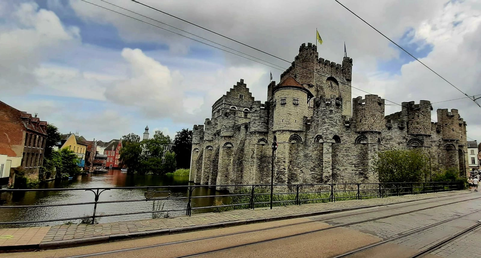 Un castello medievale in pietra con torri rotonde e merlature si trova accanto a un canale sotto un cielo parzialmente nuvoloso, con binari del tram ed edifici in primo piano e sullo sfondo.