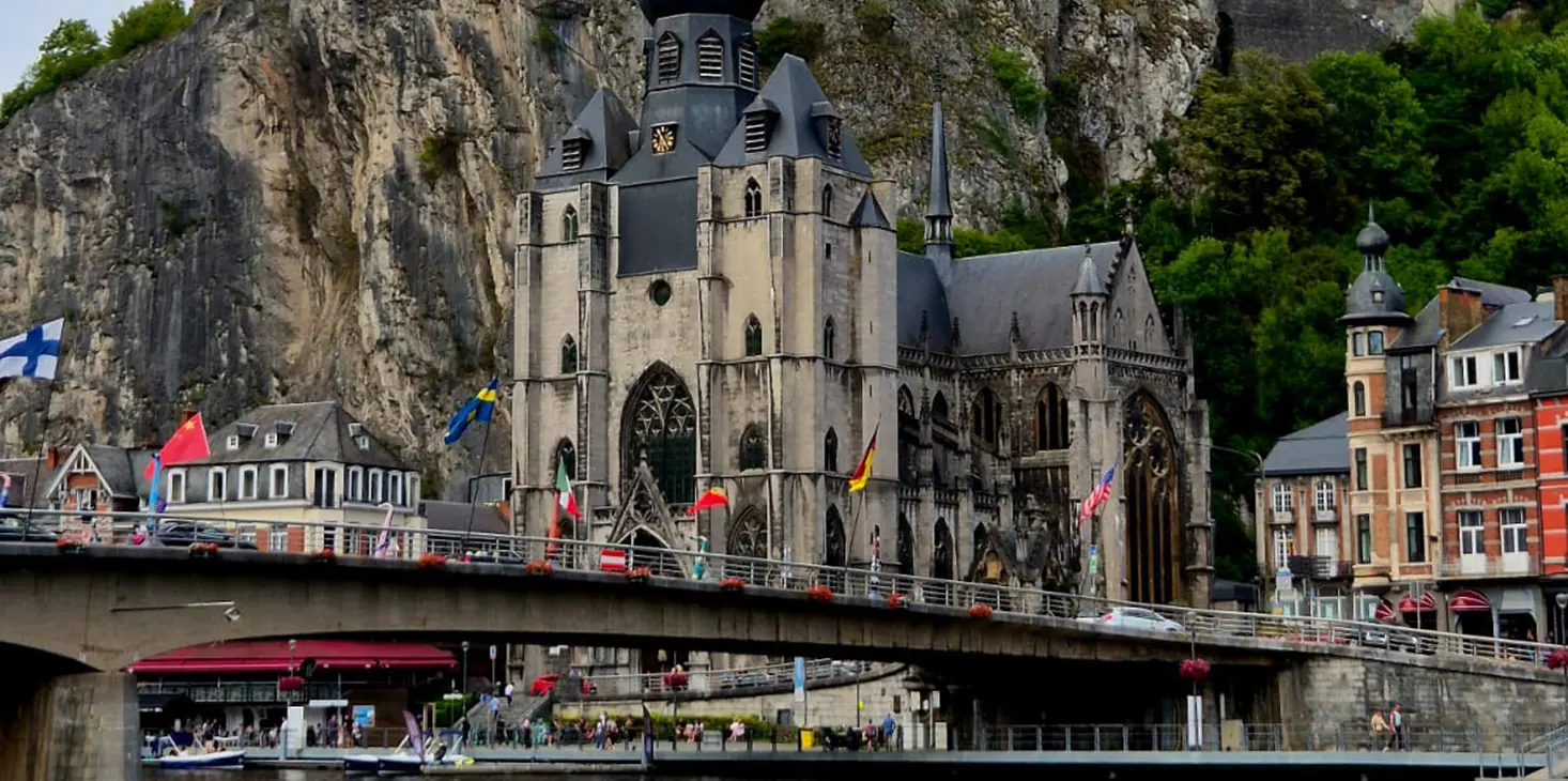 Una grande chiesa in stile gotico con un alto campanile si erge su una scogliera rocciosa, con un ponte e diverse bandiere in primo piano ed edifici colorati ai lati.