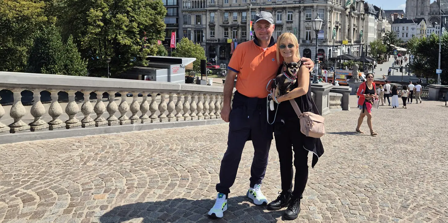Un uomo e una donna stanno insieme su un ponte acciottolato e sorridono alla telecamera. L'uomo indossa una camicia arancione e un berretto, la donna è vestita di nero e indossa occhiali da sole. Sullo sfondo si vedono edifici urbani e alberi in una giornata di sole.  