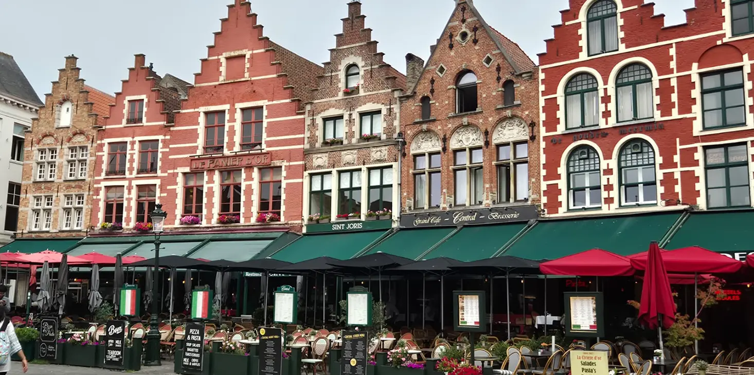 Edifici storici colorati con frontoni a gradini fiancheggiano una strada di Bruges, in Belgio. Tra questi ci sono ristoranti con verande verdi e rosse che creano un'atmosfera vivace e invitante con posti a sedere all'aperto e cartelli con i menu. 