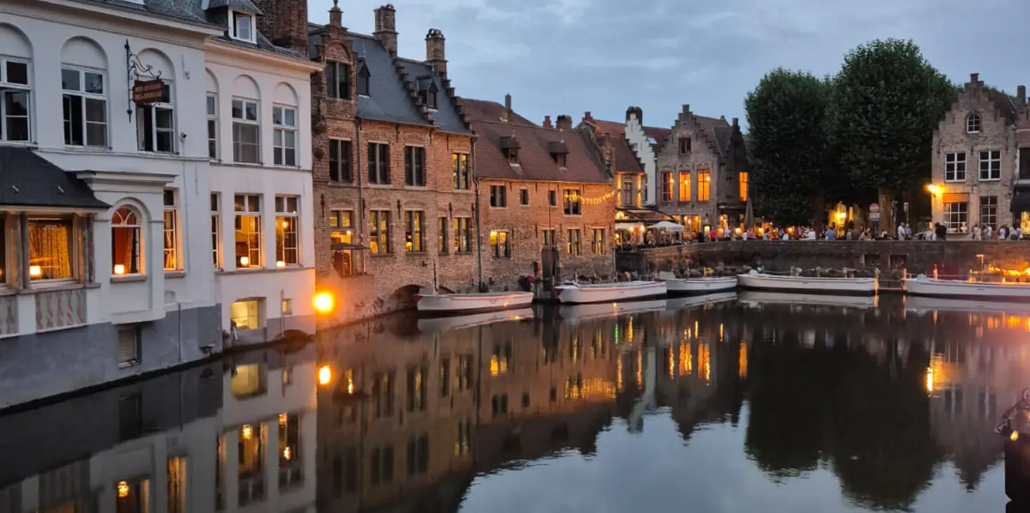 Un canale fiancheggiato da edifici storici in mattoni, caldamente illuminato al crepuscolo, le cui luci si riflettono nell'acqua calma. Sulla destra si trovano aree per sedersi all'aperto e alberi, che creano un tranquillo paesaggio urbano europeo. 