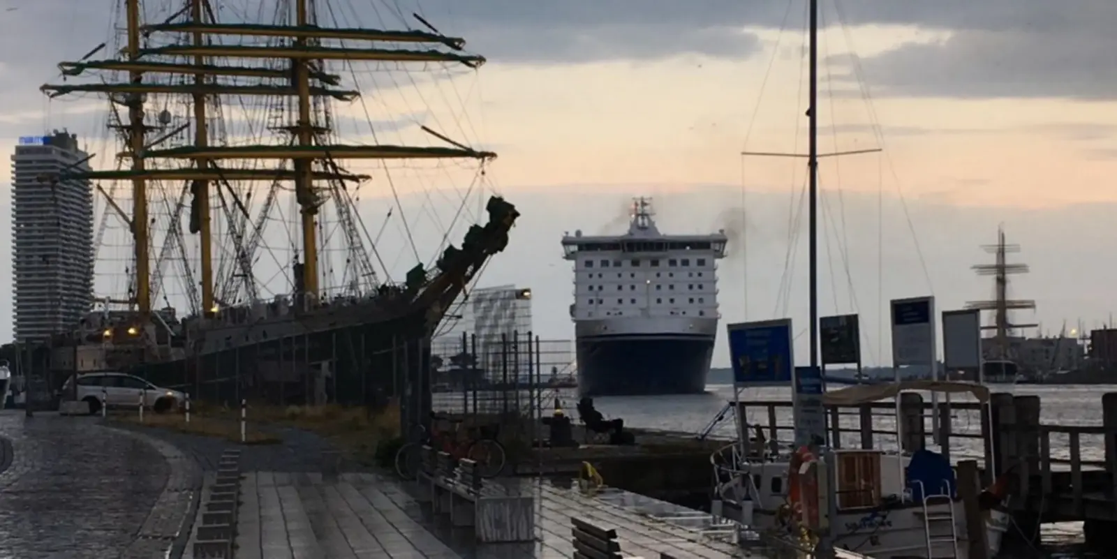 A large cruise ship approaches a harbour at dusk. A large sailing ship is docked on the left, modern buildings can be seen in the background and the sky is cloudy.