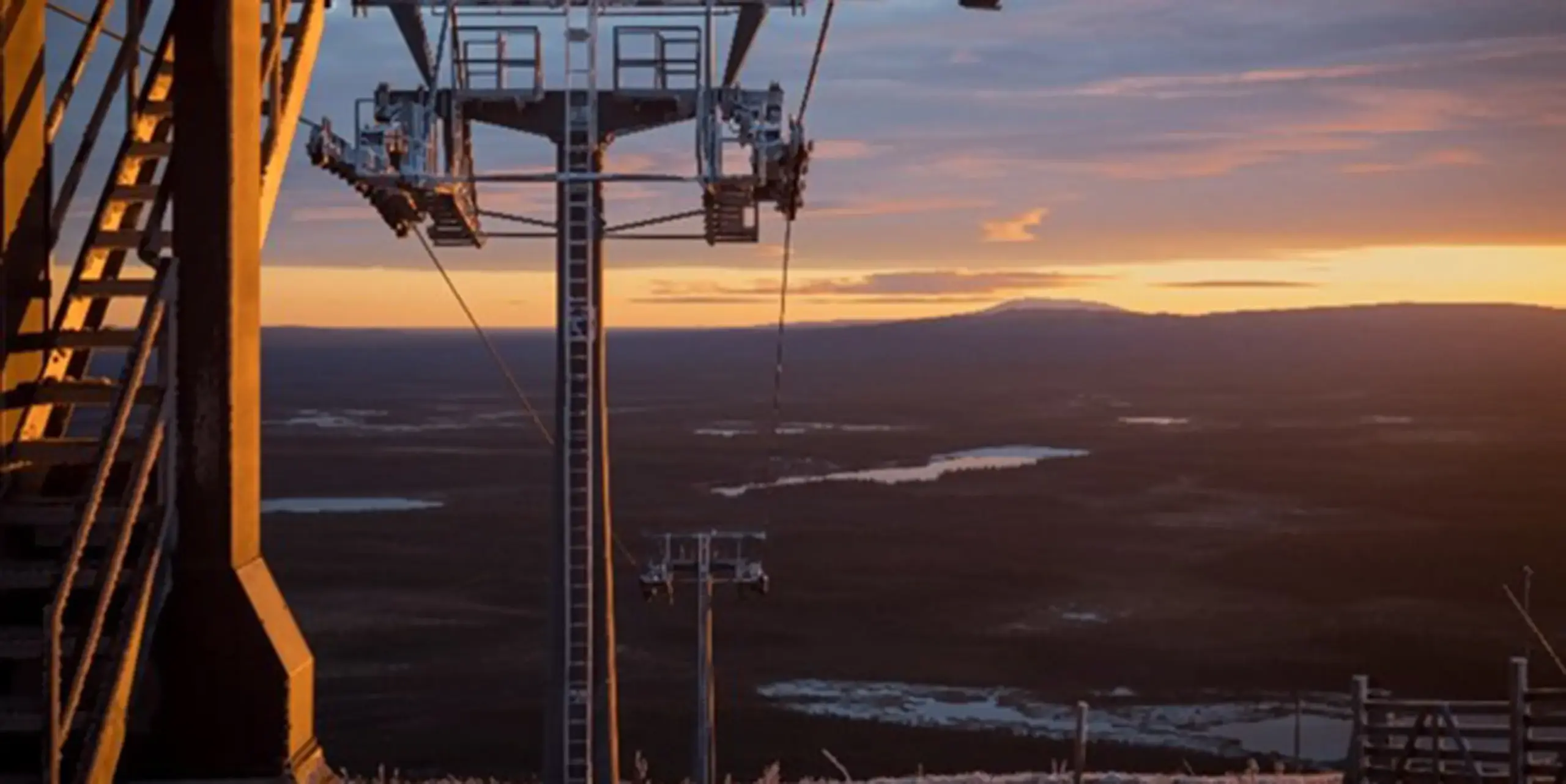A ski lift tower stands on a mountain at sunset, overlooking a vast landscape of rivers, forests and distant hills under a colourful sky.