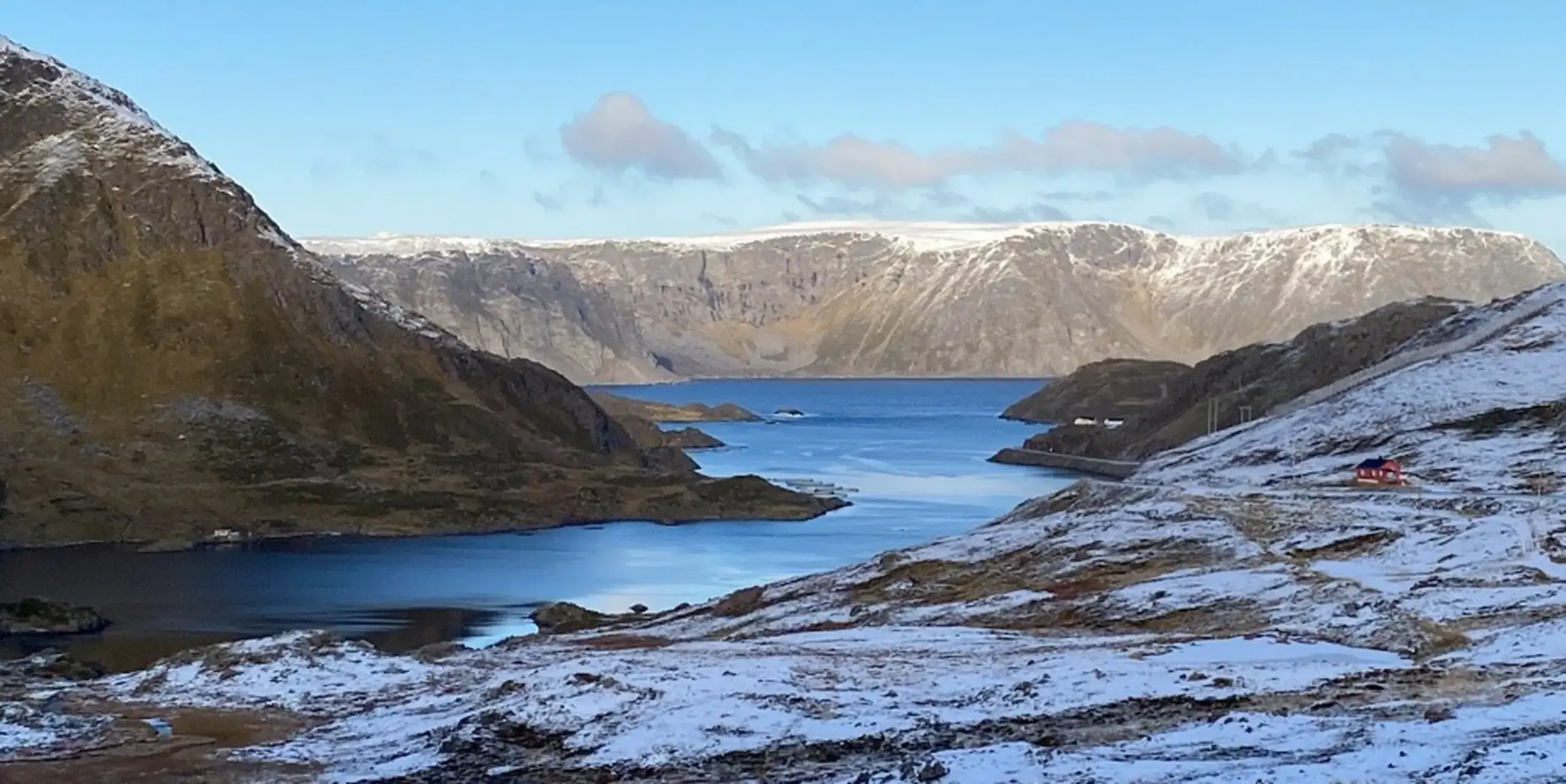 A picturesque fjord with calm, blue water, surrounded by snow-capped mountains under a clear sky, with a small red house on the right-hand slope.