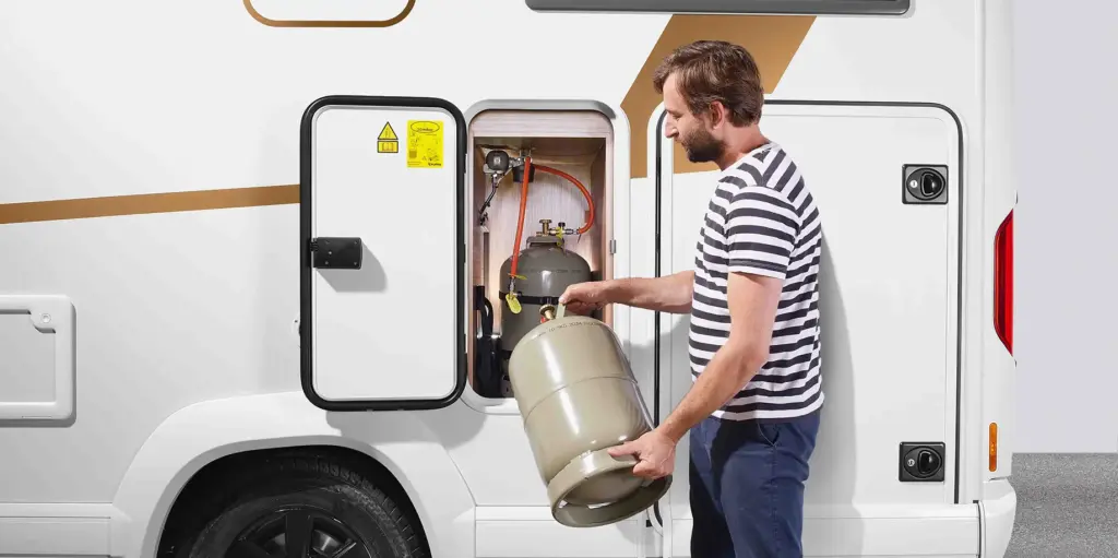 Ein Mann in einem gestreiften Hemd setzt eine Propangasflasche in ein offenes Fach an der Seite eines weißen Wohnmobils ein und bereitet sich so auf die Gasprüfung im Wohnmobil vor. Das Fach enthält eine weitere Flasche und ist mit Warnhinweisen versehen.