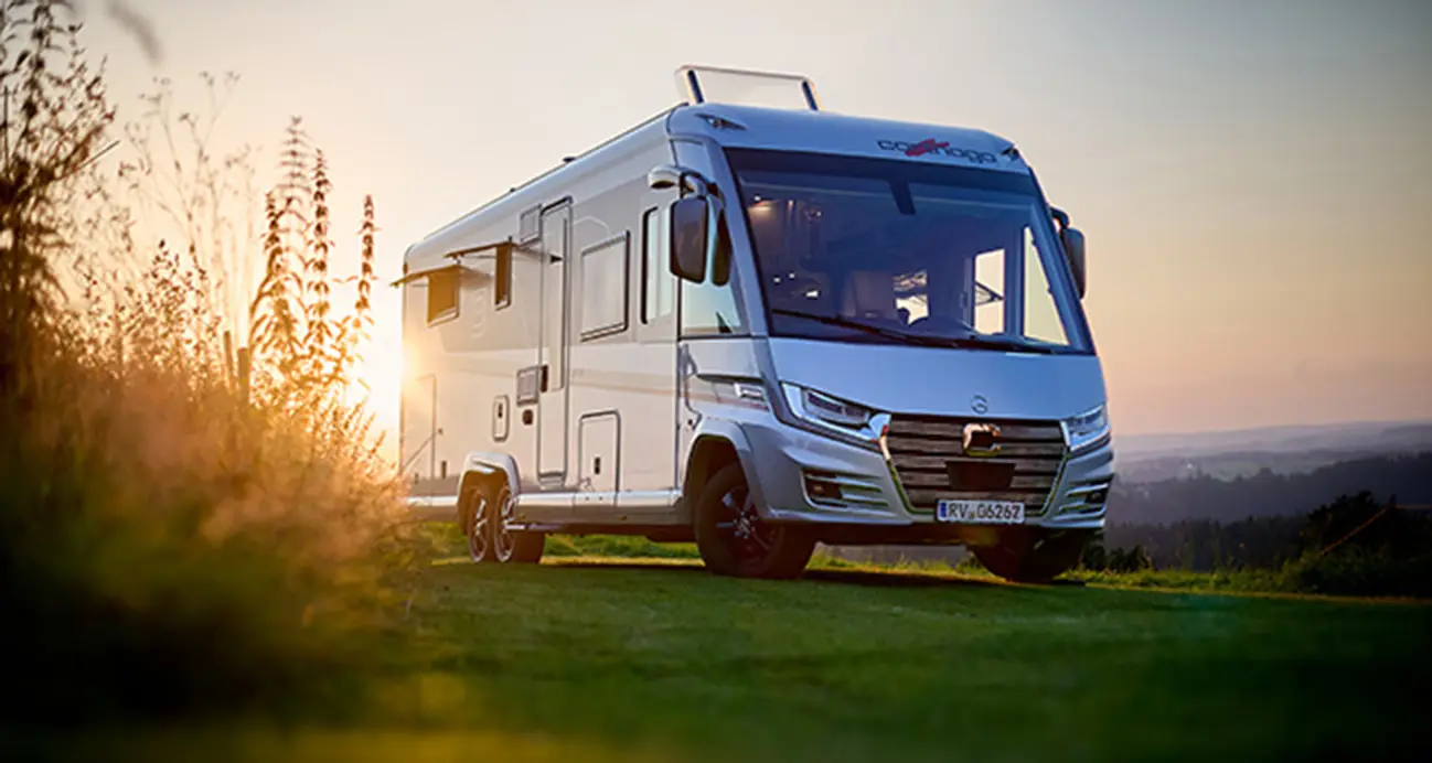Un camping-car blanc, parfait pour les longs trajets en camping-car, est garé sur une colline herbeuse au coucher du soleil. La lumière du soleil passe à travers ses baies et éclaire les hautes herbes du premier plan sous un ciel clair et doré.