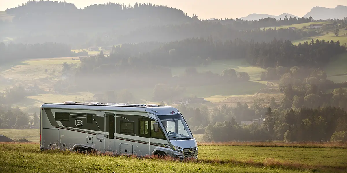 Un camping-car moderne, parfait pour les longs trajets en camping-car, se trouve dans une prairie herbeuse avec de douces collines, des forêts et des montagnes lointaines en arrière-plan, sous un ciel brumeux.
