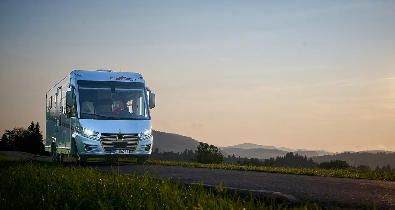 Un camping-car blanc roule le long d'une route de campagne au coucher du soleil, parfait pour les longs trajets en camping-car, avec de l'herbe verte au premier plan et des collines et des arbres en pente douce au loin sous un ciel dégagé.