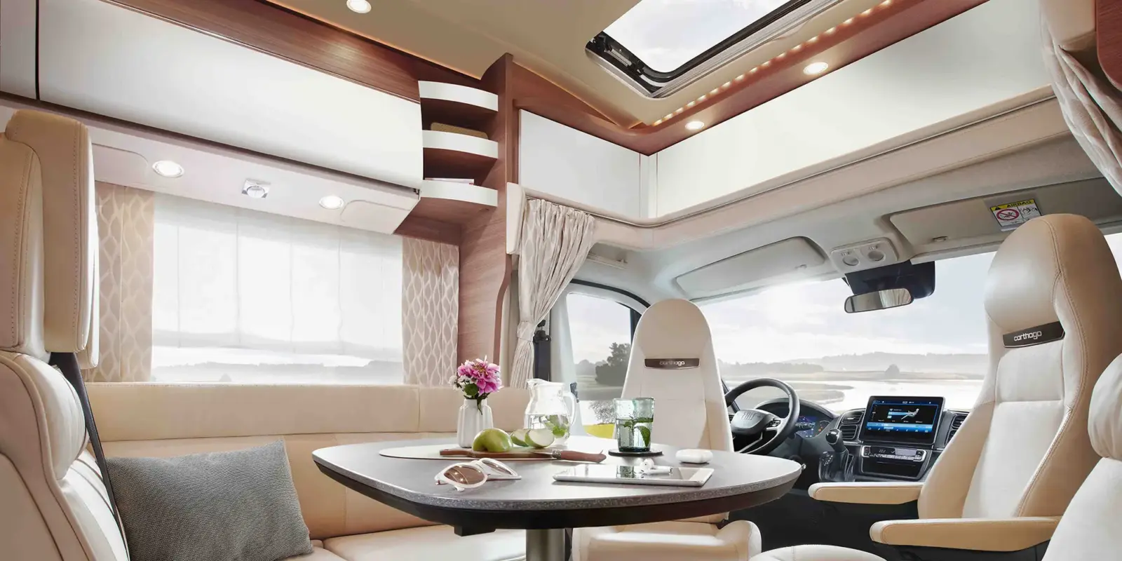 Modernes Wohnmobil-Interieur des Carthago Chic C Line T mit cremefarbenen Sitzgelegenheiten, einem runden Esstisch mit Geschirr, Getränken und Obst, großen Fenstern für natürliches Licht und einer Panorama-Windschutzscheibe, die den Blick nach draußen freigibt.